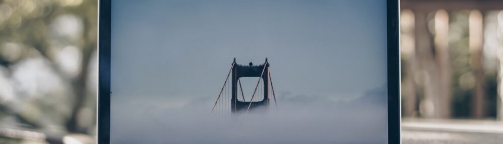A laptop displaying a foggy bridge on its screen, placed on a wooden table in a natural setting.