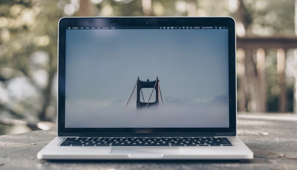 A laptop displaying a foggy bridge on its screen, placed on a wooden table in a natural setting.